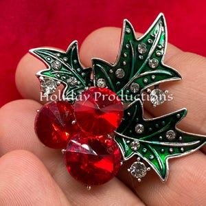 May include: A silver-toned brooch with red and green enamel. The brooch features three red rhinestones and two green leaves with white rhinestones. The leaves are outlined in silver.