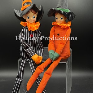 May include: Two posable dolls with pointed hats and ruffled collars. One doll wears a black and white striped suit with an orange hat, while the other wears an orange textured suit with a black hat. The text Holiday Productions is visible.