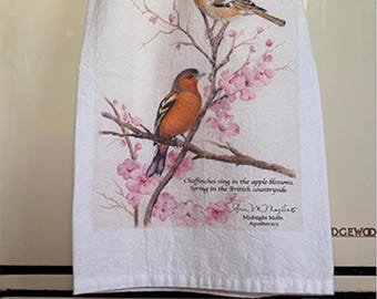 May include: A white tea towel with a watercolor illustration of two birds perched on a branch with pink blossoms. The text reads "Chaffinches sing in the apple blossoms, Spring in the British countryside." and is signed "Sue Magill, Midnight Moon Apothecary."