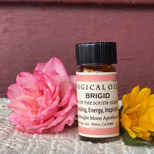 May include: A small brown glass bottle with a black cap, labeled "Magical Oil Brigid of the South-Element Healing, Energy, Inspiration Midnight Moon Apothecary Box 1115 Willits, CA 95490 1.0 fl oz". The bottle is sitting on a white crocheted surface with a pink rose and a yellow flower.