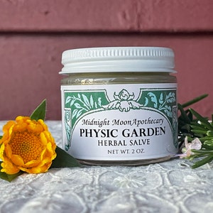 May include: A white jar with a green label that reads "Midnight Moon Apothecary Physic Garden Herbal Salve Net Wt. 2 oz."