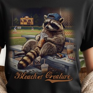 Può includere: T-shirt nera con l'illustrazione di un procione con occhiali da sole e berretto da baseball, che beve da una tazza con una cannuccia. Il testo "Bleacher Creature" è sotto il procione. Lo sfondo mostra un campo da baseball.