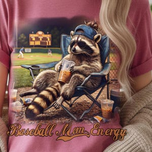 Può includere: Una maglietta rosa polveroso presenta un procione con occhiali da sole e un berretto da baseball, che si rilassa su una sedia pieghevole blu mentre beve caffè freddo. La maglietta ha il testo "Baseball Mom Energy" in oro.