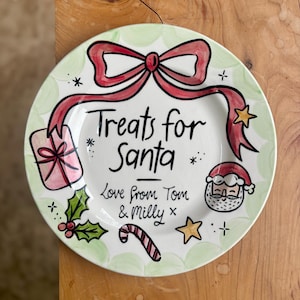 May include: A hand-painted ceramic plate with a Christmas theme. The plate features a red bow, the text "Treats for Santa", and illustrations of a gift, holly, a candy cane, and Santa Claus. The plate is trimmed with a green border.