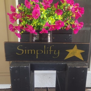 May include: A black wooden plant stand with a sign that reads "Simplify" in yellow script, and a yellow star. The stand holds a pot of vibrant pink flowers with green foliage.
