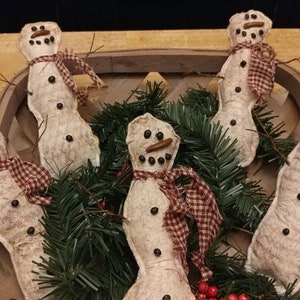 May include: Five handmade fabric snowman ornaments with brown eyes, black buttons, and red and white gingham scarves. The snowmen are arranged in a basket with green pine branches and red berries.