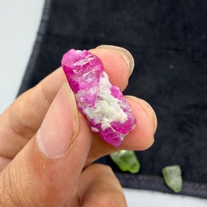 May include: A raw, uncut ruby crystal with a vibrant pink hue and a rough, textured surface. The ruby has areas of white and is held in a hand. Two green crystals are in the background.