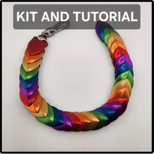 Op de afbeelding: Een regenboogkleurige schubbenketting met overlappende metalen schubben in rood, oranje, goud, groen, blauw en paars. De ketting is verbonden met een zilverkleurige metalen karabijnhaak. De tekst KIT AND TUTORIAL is bovenaan zichtbaar.