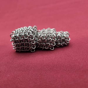 May include: A silver-colored chainmail sculpture composed of three interconnected cubes of varying sizes. The structure is crafted from numerous small, interlocking metal rings, creating a textured, grid-like pattern across each cubic surface.