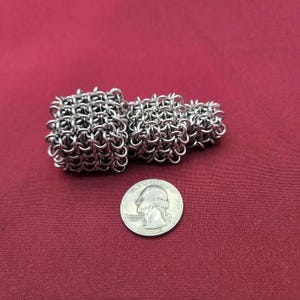 May include: A silver-colored chainmail object composed of three interconnected, tiered square segments of varying sizes. The object is positioned next to a United States quarter coin, which has a diameter of approximately 0.955 inches for scale.