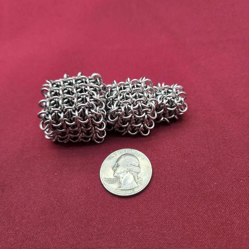 May include: A silver-colored chainmail object composed of three interconnected, tiered square segments of varying sizes. The object is positioned next to a United States quarter coin, which has a diameter of approximately 0.955 inches for scale.