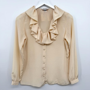 May include: A long-sleeved, light peach-colored blouse featuring a deep V-neckline adorned with a wide, ruffled collar. The fabric has a subtle, tonal dotted pattern and the front is secured with a vertical row of matching fabric-covered buttons.