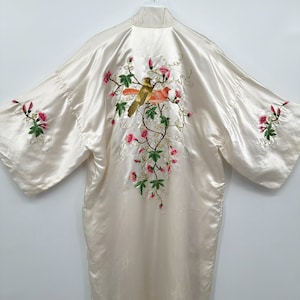 May include: A cream-colored satin robe featuring intricate embroidery on the back and sleeves. The back design displays two birds perched on flowering branches with pink blossoms and green leaves. Similar floral embroidery appears on each sleeve.