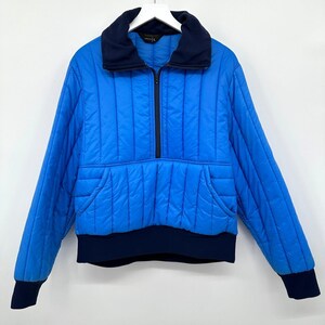 May include: A blue quilted jacket with a navy blue collar, cuffs, and hem. The jacket has a half-zip closure and a front pocket. The jacket is hanging on a white hanger against a white background.