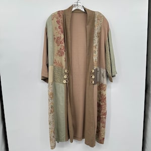 May include: A long, open-front cardigan in shades of tan, brown, and green, with floral patchwork panels. The cardigan features three sets of gold-colored buttons and is hanging on a silver hanger.