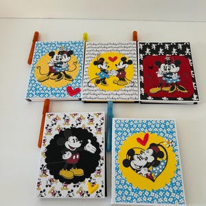 May include: Five rectangular notebooks featuring Mickey and Minnie Mouse illustrations. The covers display various patterns, including blue and white flowers, black and white motifs, and yellow scalloped circles with heart accents and text.