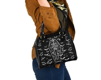 May include: Black handbag with a gothic design featuring a skeleton in a coffin and bat silhouettes. The bag has a glossy finish, black handles, and gold-colored hardware. The bag is carried by a person wearing a brown jacket and blue jeans.