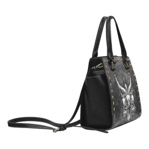 May include: Black handbag with a white graphic print of an angel with wings. The bag has gold-coloured studs and a detachable shoulder strap.
