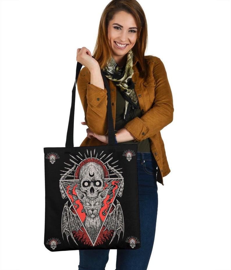May include: Black tote bag featuring a graphic print of a skull with a cat face inside, surrounded by flames and a triangle. The skull has a third eye and a crescent moon on its forehead. There are four small skulls around the main graphic.