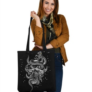 Op de afbeelding: Een zwarte tote bag met een witte afbeelding van een schedel met een slang eromheen gewikkeld. De slang heeft een geel oog. De tas is versierd met witte stippen.