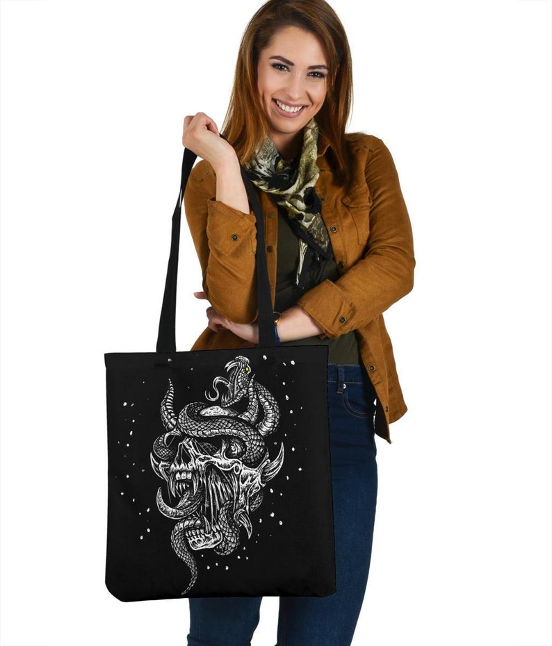 Op de afbeelding: Een zwarte tote bag met een witte afbeelding van een schedel met een slang eromheen gewikkeld. De slang heeft een geel oog. De tas is versierd met witte stippen.