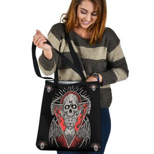 May include: Black tote bag featuring a graphic of a skull and a cat with wings within a triangle. The design is in red, white, and black. Smaller skulls are depicted around the edge of the bag.