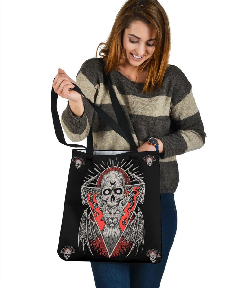 May include: Black tote bag featuring a graphic of a skull and a cat with wings within a triangle. The design is in red, white, and black. Smaller skulls are depicted around the edge of the bag.