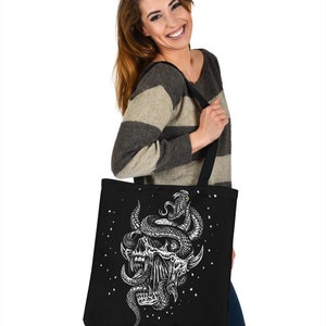 Op de afbeelding: Zwarte tote bag met een witte afbeelding van een schedel met een slang eromheen gewikkeld. De slang heeft een geel oog. De achtergrond is zwart met witte stippen.