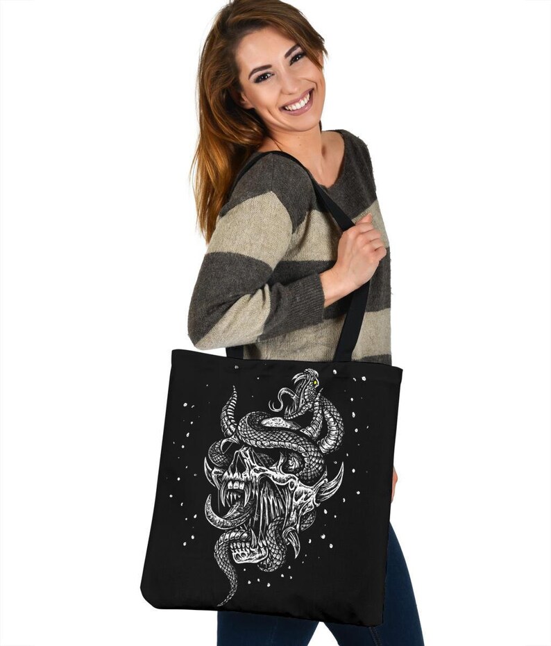 Op de afbeelding: Zwarte tote bag met een witte afbeelding van een schedel met een slang eromheen gewikkeld. De slang heeft een geel oog. De achtergrond is zwart met witte stippen.