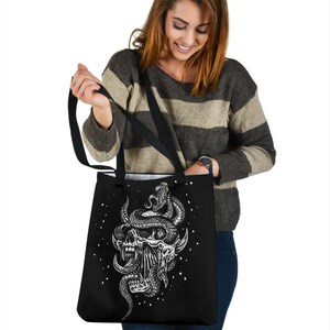 Op de afbeelding: Zwarte tote bag met een witte afbeelding van een schedel met een slang eromheen gewikkeld. De slang heeft een wit oog en de schedel heeft een witte mond met tanden.
