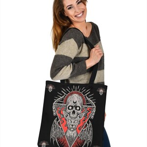 May include: A black tote bag featuring a graphic print of a skull with a crescent moon, a winged cat, and flames. The design is in a triangular shape with skulls at the corners.