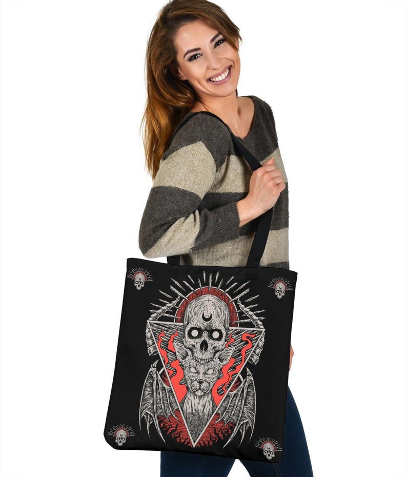 May include: A black tote bag featuring a graphic print of a skull with a crescent moon, a winged cat, and flames. The design is in a triangular shape with skulls at the corners.