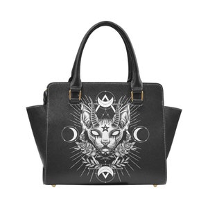 Op de afbeelding: Zwarte leren handtas met een witte grafische print van een kat met hoorns en een pentagram. De afbeelding is omringd door een cirkel van manen en een ster.
