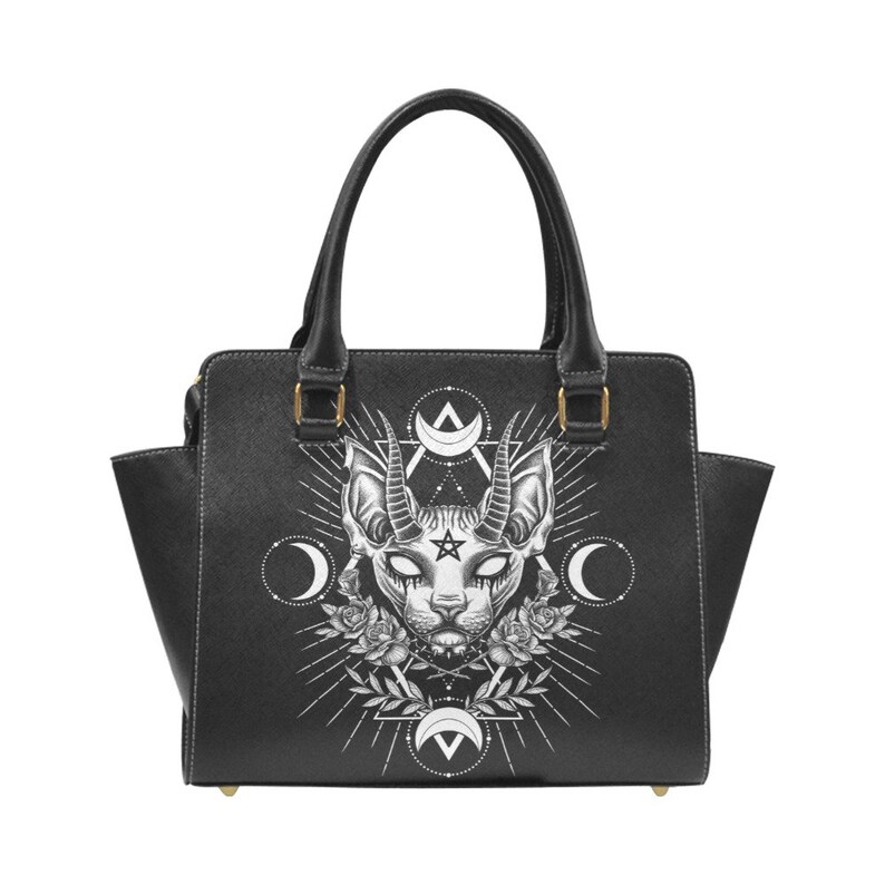Op de afbeelding: Zwarte leren handtas met een witte grafische print van een kat met hoorns en een pentagram. De afbeelding is omringd door een cirkel van manen en een ster.