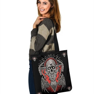May include: A black tote bag featuring a skull and wings design. The skull is surrounded by flames and a crescent moon. The bag has a black strap and is being held by a person.