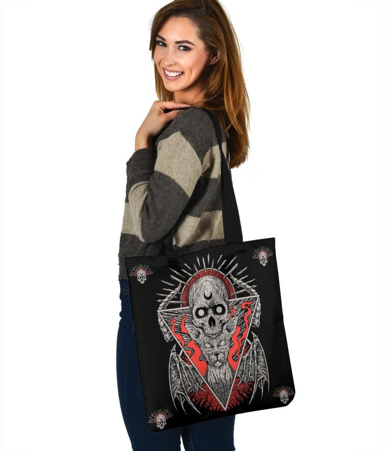 May include: A black tote bag featuring a skull and wings design. The skull is surrounded by flames and a crescent moon. The bag has a black strap and is being held by a person.
