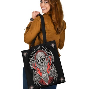 May include: Black tote bag featuring a graphic of a skull with wings and a crescent moon within a triangle. The skull is surrounded by flames, and the background is black with small skulls scattered around the edges.