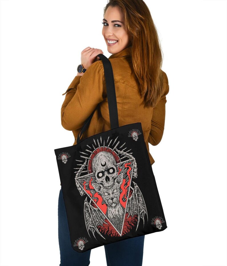 May include: Black tote bag featuring a graphic of a skull with wings and a crescent moon within a triangle. The skull is surrounded by flames, and the background is black with small skulls scattered around the edges.