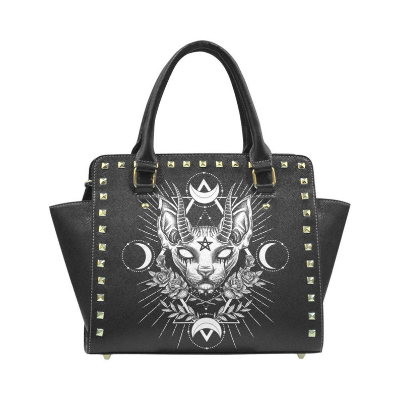 Op de afbeelding: Zwarte handtas met een witte afbeelding van een kat met hoorns en een pentagram. De tas heeft gouden studs rond de randen.