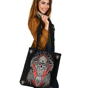 May include: Black tote bag featuring a skull and flames design. The skull has a crescent moon on its forehead and is surrounded by a triangle. The flames are red and orange. The bag has black straps and a black lining.