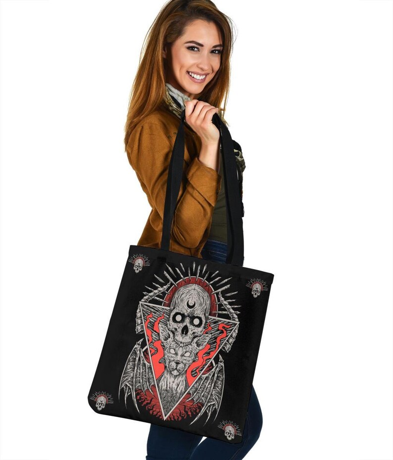 May include: Black tote bag featuring a skull and flames design. The skull has a crescent moon on its forehead and is surrounded by a triangle. The flames are red and orange. The bag has black straps and a black lining.