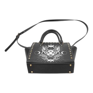Op de afbeelding: Zwarte leren handtas met een witte afbeelding van een kat met hoorns en een pentagram. De tas heeft gouden studs en een schouderband.
