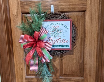 May include: A Christmas wreath with a red and white striped bow and green pine branches. The wreath has a sign that says "Meet me under the mistletoe Kisses Guaranteed".