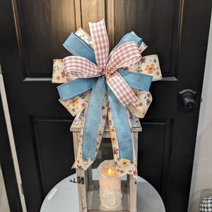 May include: A white lantern with a floral patterned ribbon bow on top. The lantern has a glass panel and a candle inside. The candle is lit and glowing.