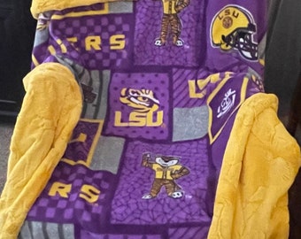 May include: Purple and gold fleece blanket with a patchwork design featuring the LSU logo and mascot. The blanket has a yellow fleece backing.