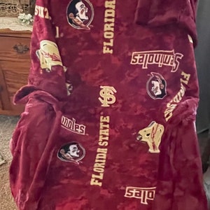 May include: A maroon and gold fleece blanket featuring the words "Florida State" and "Seminoles" in gold lettering. The blanket has a repeating pattern of the Seminoles logo and is draped over a chair.