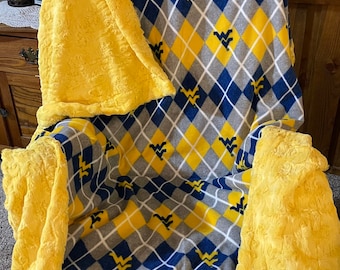 May include: A yellow and blue fleece blanket with a diamond pattern and a team logo. The blanket is folded and displayed on a chair.