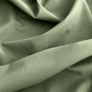 May include: Close-up of olive green fabric with soft folds and subtle texture. The fabric appears to be a lightweight material, possibly cotton or linen, with a slight sheen. The folds create shadows and highlights, adding depth to the image.