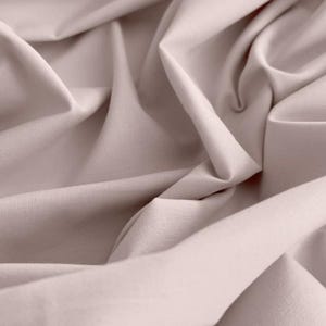 May include: A close-up shot of a light mauve fabric with gentle folds and shadows. The material looks to be a smooth, woven textile, perhaps linen or cotton, with a matte finish. The image emphasises the texture and the way the fabric drapes.
