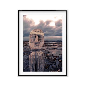 May include: A wooden sculpture of a face with a white cloth blindfold, standing on a rocky beach with a cloudy sky in the background.